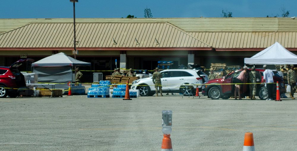 A supply pickup site after Hurricane Ian: tents, pallets of water and guardsmen loading cars in a shopping center lot.